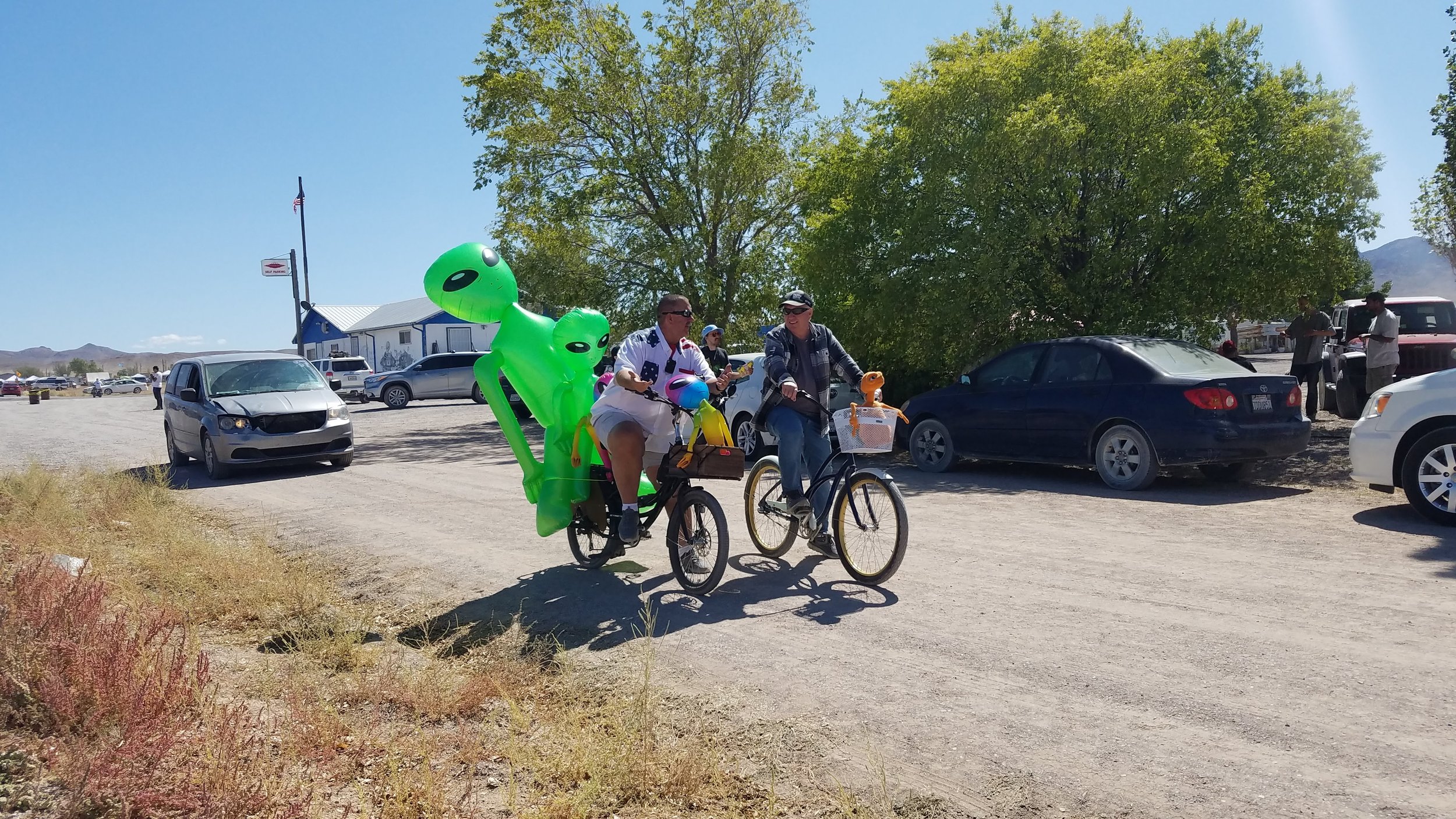 Two men riding bikes with inflatable alien decorations attached to their bikes, in a parking area of the Little A'Le'Inn in Rachel, Nevada, with cars, trees, and a building in the background.