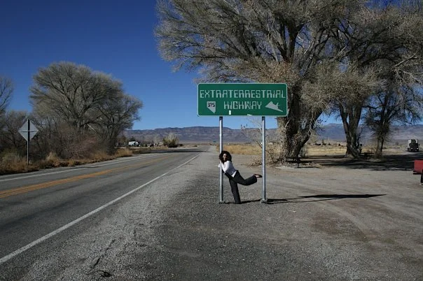 Aleza under a sign that reads 'Extraterrestrial Highway,' situated by the side of a deserted road in a desert landscape with large trees and mountains in the background.