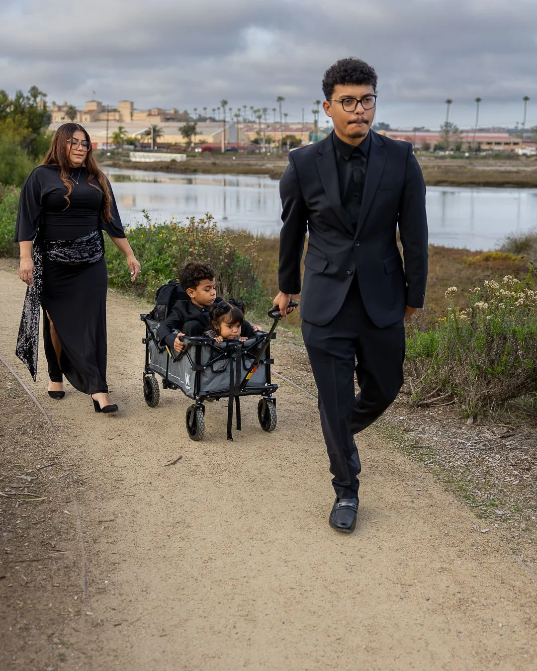 A family walking outdoors on a dirt path near a body of water with buildings and palm trees in the background, all dressed in formal black attire.