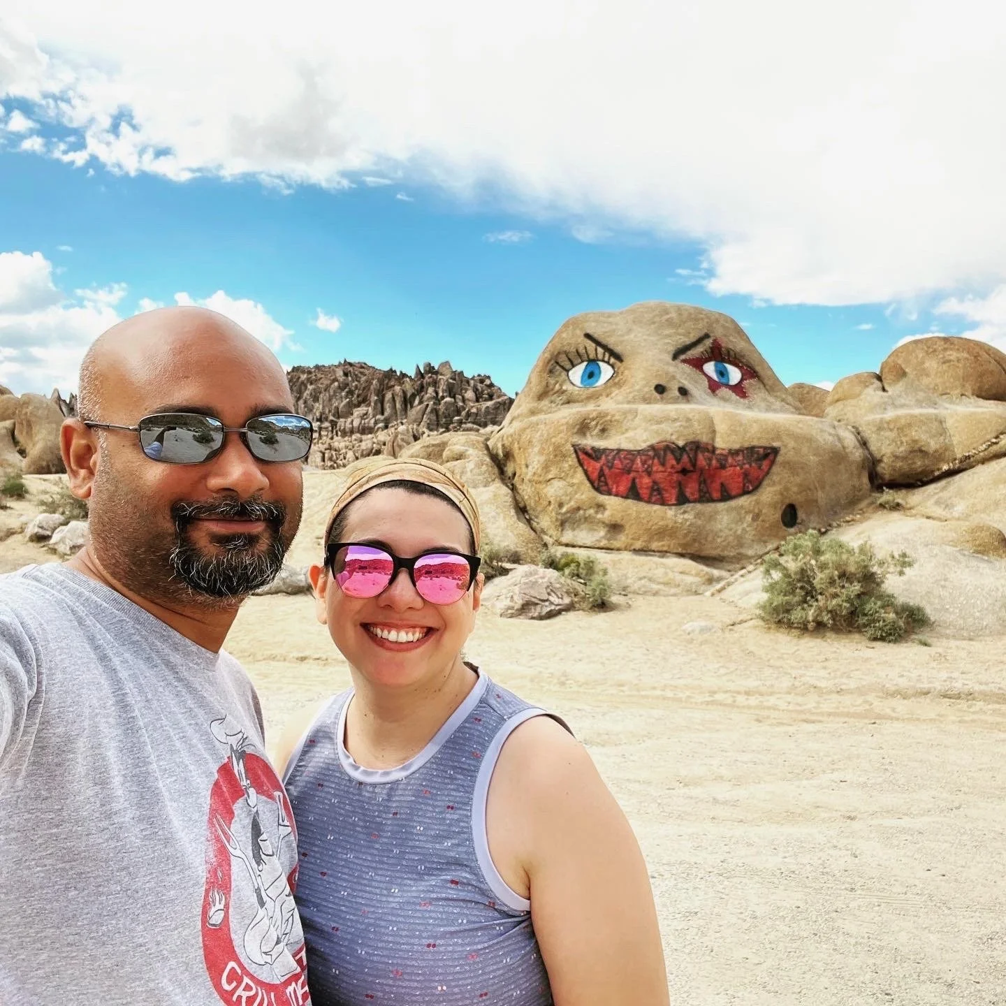 Ruwan and Diana wearing sunglasses taking a selfie in front of a large rock formation with painted cartoonish eyes, mouth, and facial features, resembling a face with a blue sky and some clouds in the background.