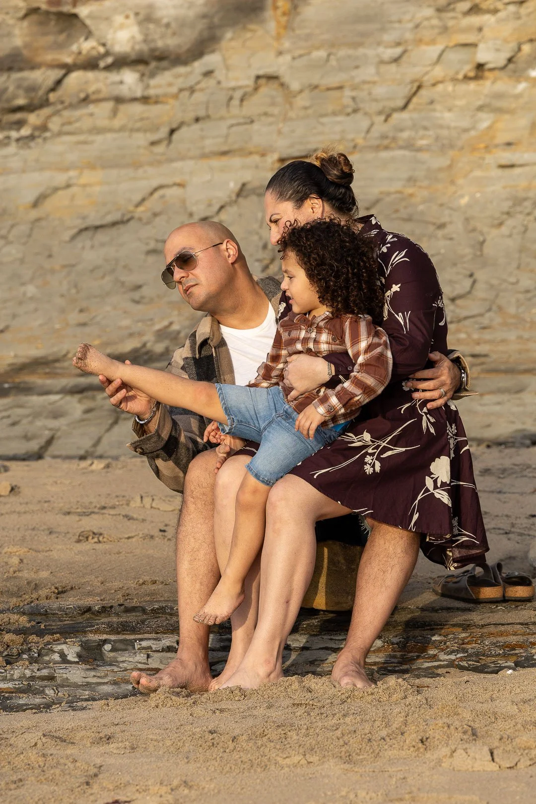 A family of three sitting on a rock at the beach, with a rocky wall in the background. The father wears sunglasses and a plaid shirt, the mother wears a dark floral dress, and the child wears a plaid shirt and denim shorts.