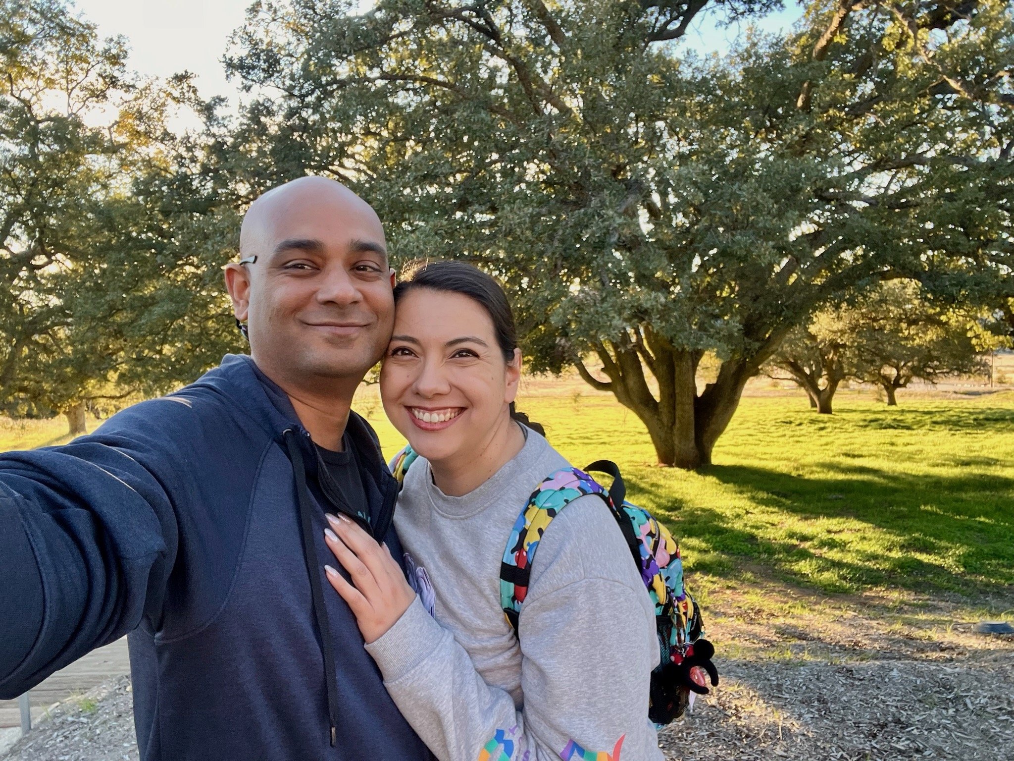 A Ruwan and Diana smiling and hugging outdoors with a tree and the sun setting in the background.