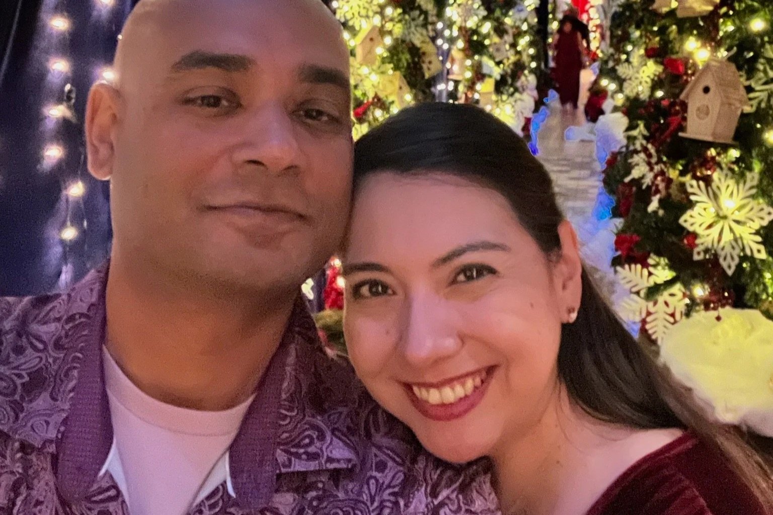 A smiling Ruwan and Diana taking a selfie in front of Christmas decorations, including a decorated Christmas tree with lights, ornaments, and a small birdhouse.