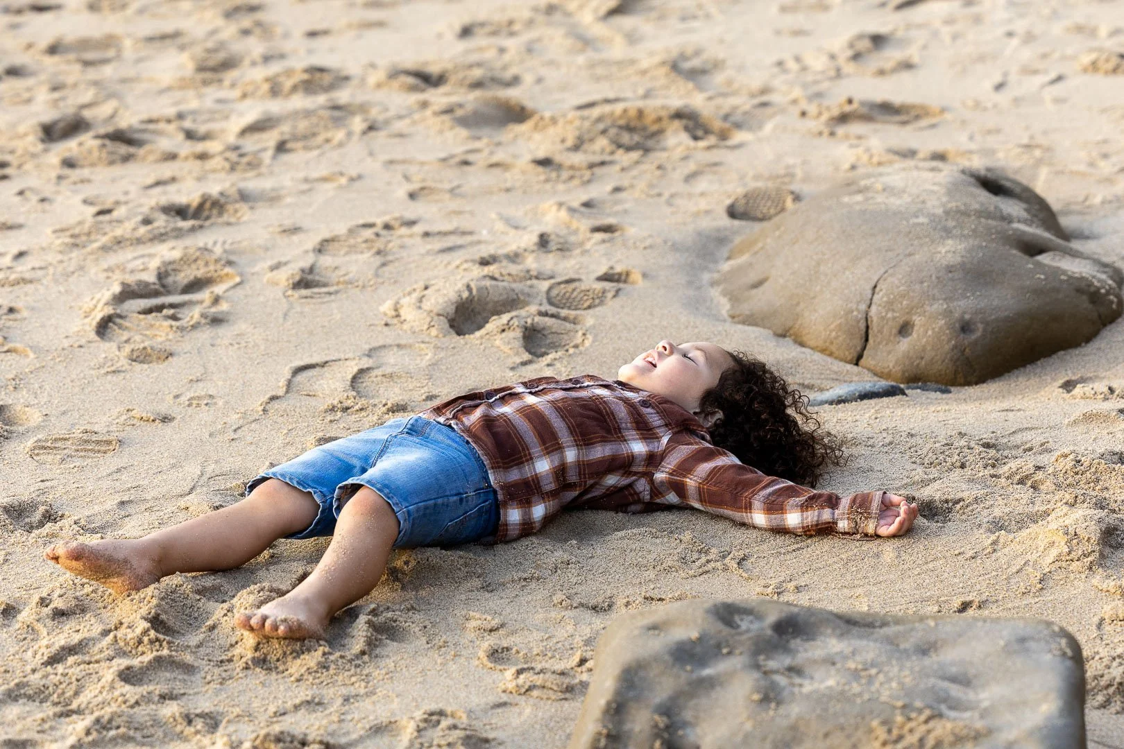 Child lying on sandy beach with eyes closed and arms outstretched making "Sand Angels", surrounded by rocks on beach