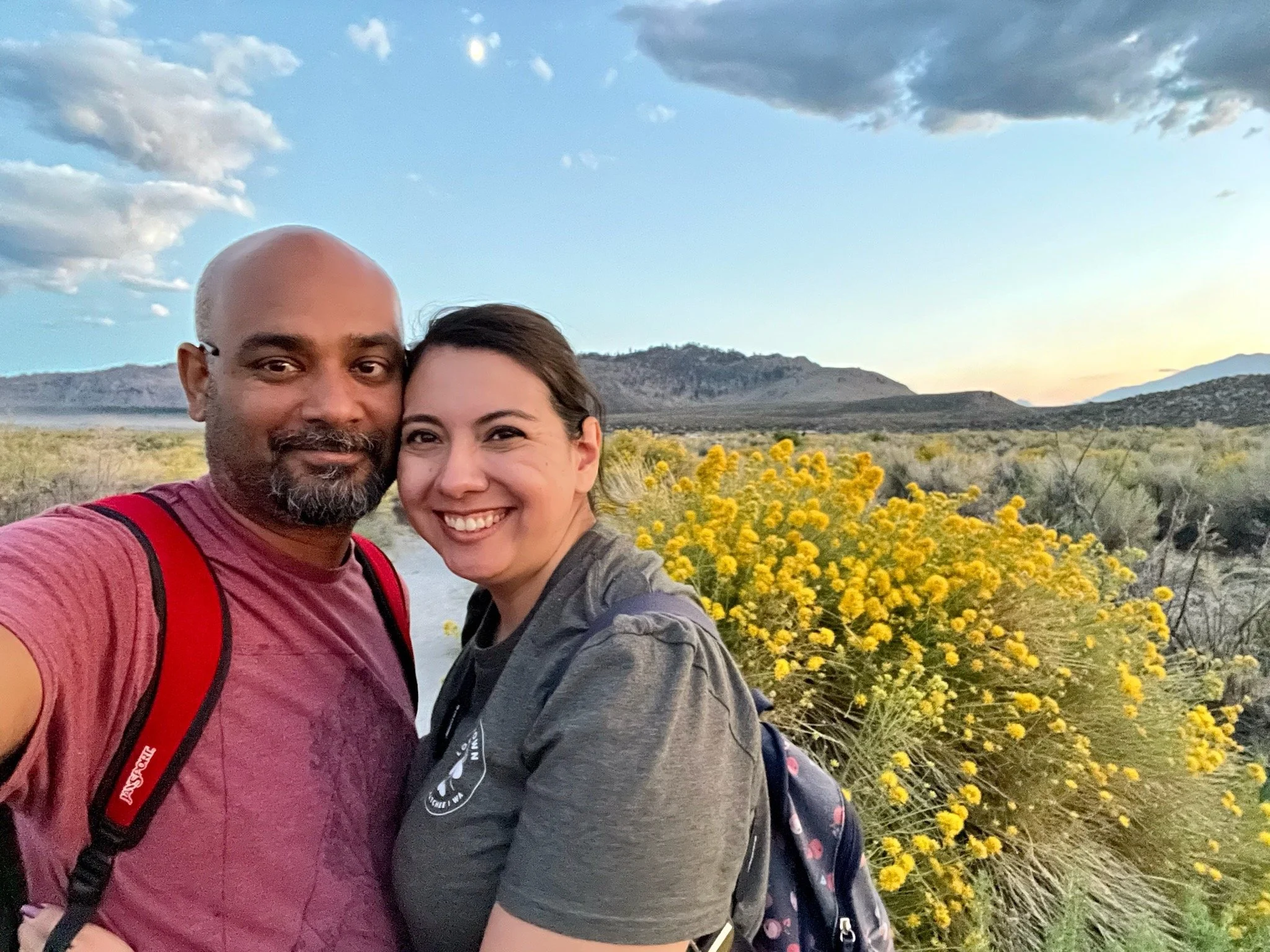 Ruwan and Diana taking a selfie outdoors during sunset in a desert landscape with yellow wildflowers, mountains, and partly cloudy sky in the background.