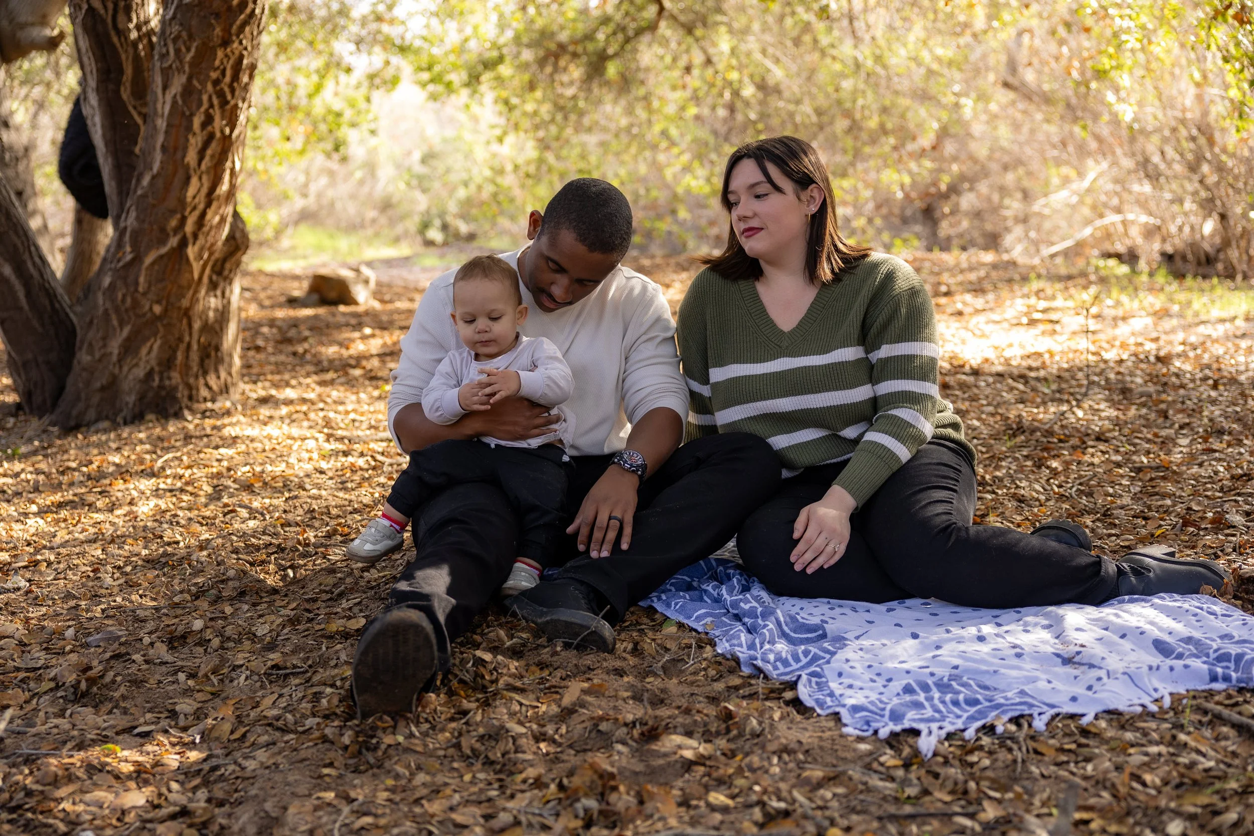 A family of three sitting on a blanket in a forest, with trees and fallen leaves around them. The man is holding a toddler, and the woman sits beside them.