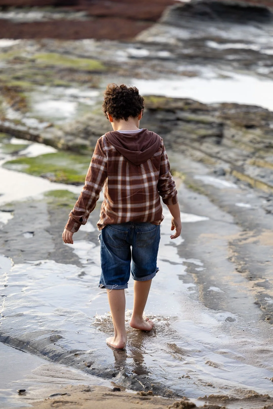 Boy walking barefoot along wet rocks and sand at the shoreline with water and tide pools.