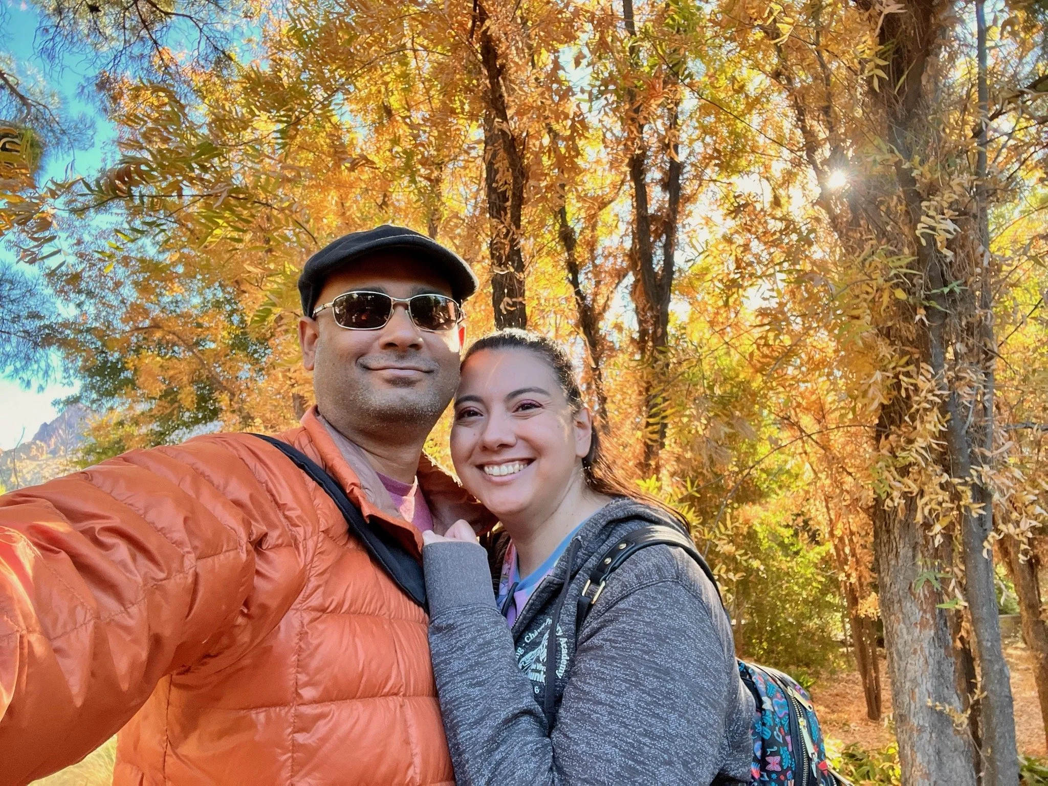 Ruwan and Diana taking a selfie outdoors during fall, surrounded by trees with colorful orange and yellow leaves.