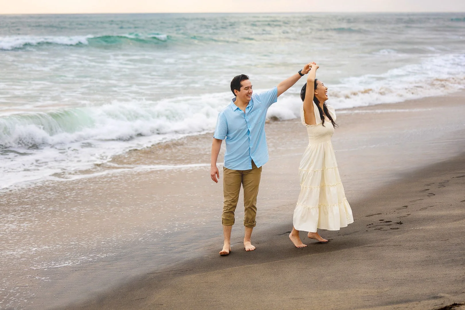 A couple holding hands and dancing on a beach with ocean waves in the background.