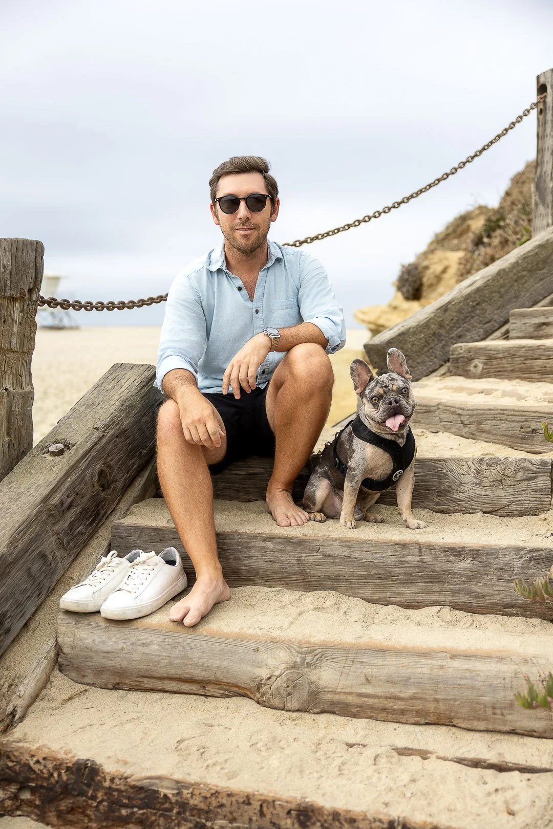 A man sitting on wooden stairs at the beach with a French Bulldog. The man is wearing sunglasses, a light blue shirt, and black shorts. Behind him, there are sandy stairs, a chain, and rocks. The man’s slippers are on the sand next to the stairs, and the bulldog is sitting beside him with its tongue out.