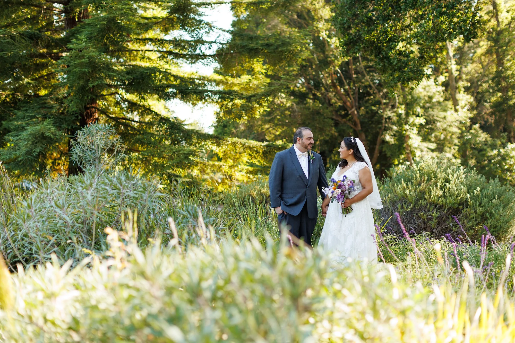 Bride and groom standing in a lush garden, smiling at each other, surrounded by greenery and flowers, during a wedding ceremony.