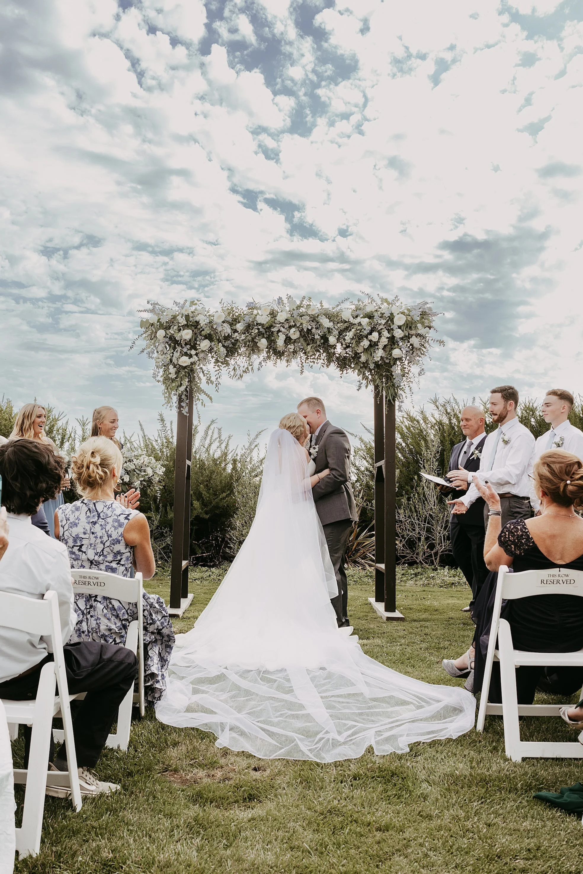 A bride and groom kissing under an outdoor wedding arch decorated with white flowers, surrounded by friends and family on a grassy lawn on a partly cloudy day.