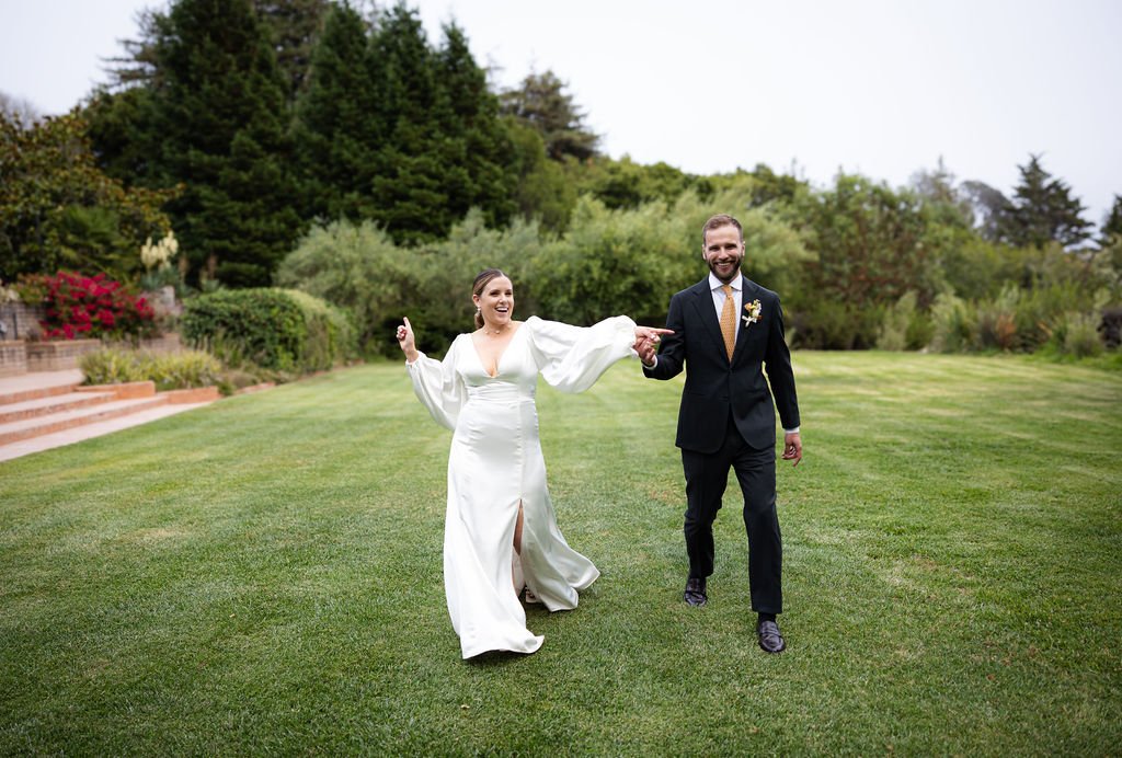 A bride and groom walking hand in hand on a green lawn, smiling, with trees and shrubs in the background.