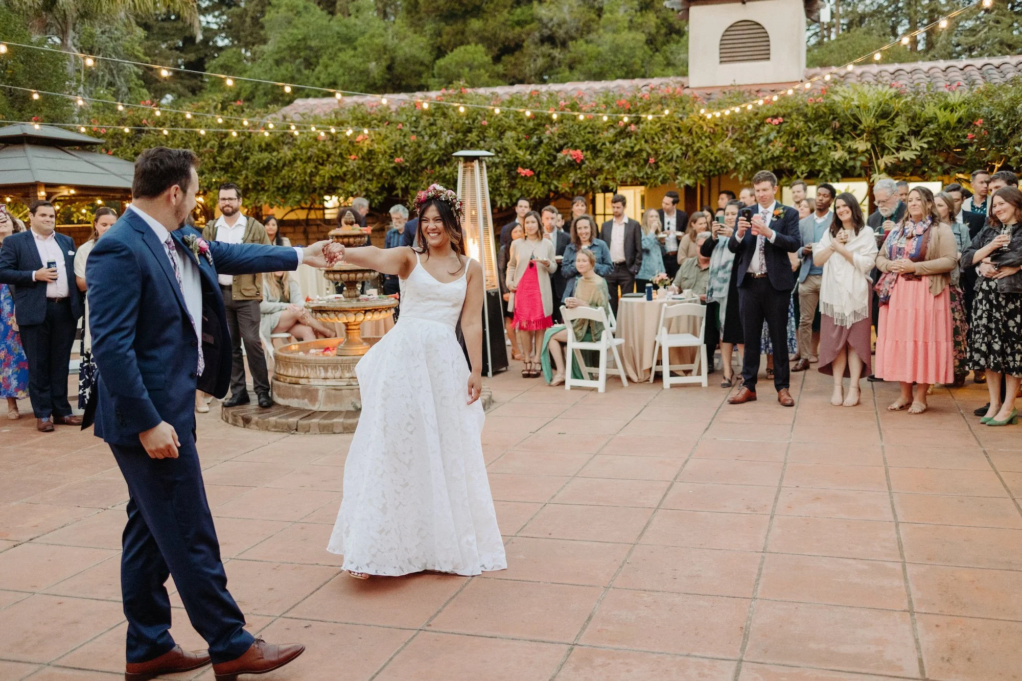 A bride and groom holding hands, dancing at their wedding reception outdoors with guests watching and taking photos. String lights are hanging above, and there are tables and lush greenery in the background.