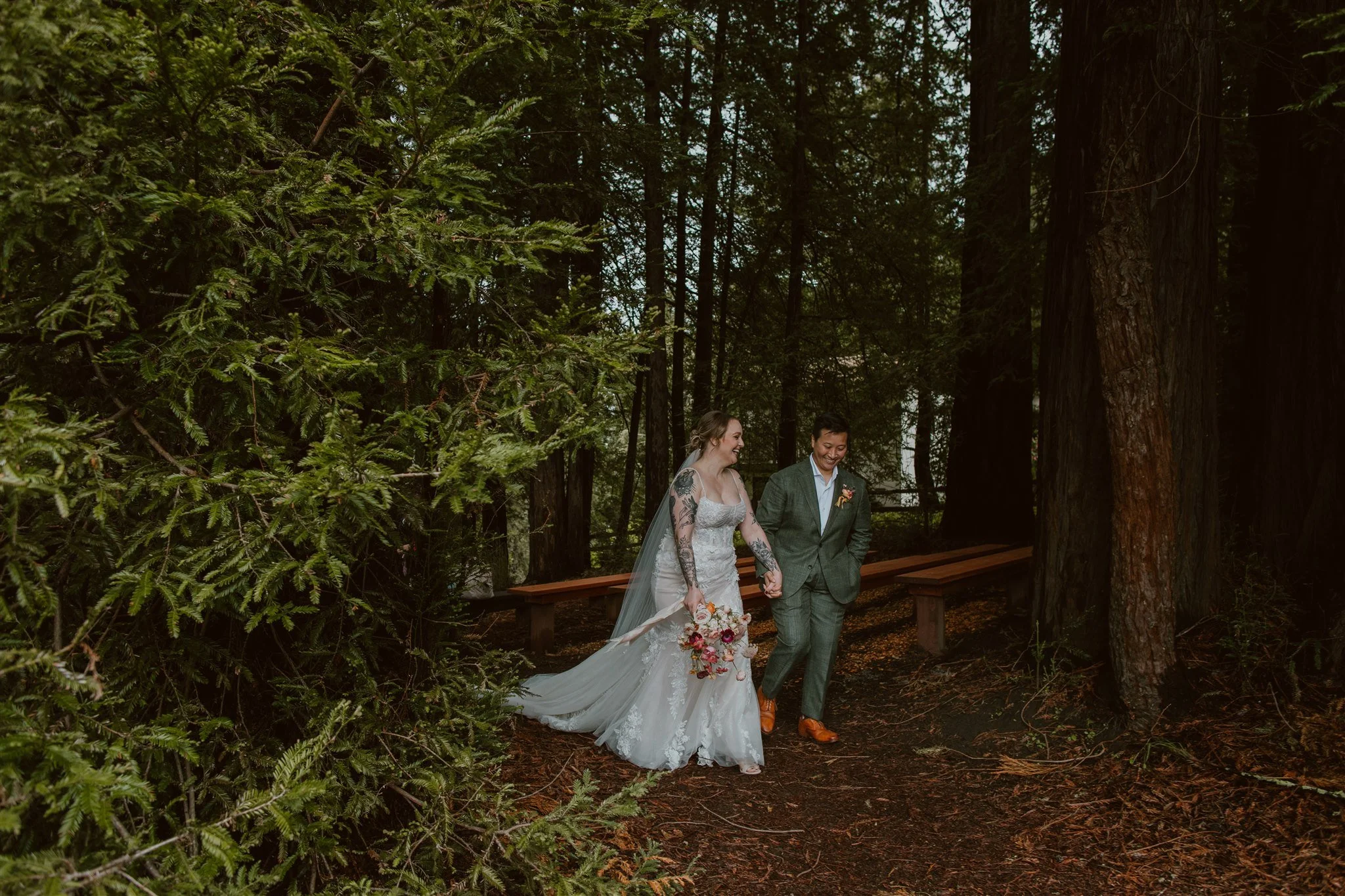 A newlywed couple walking hand in hand through a forest, with the bride in a white wedding dress and the groom in a green suit. The bride is holding a bouquet of flowers, and they are smiling at each other surrounded by tall trees and greenery.