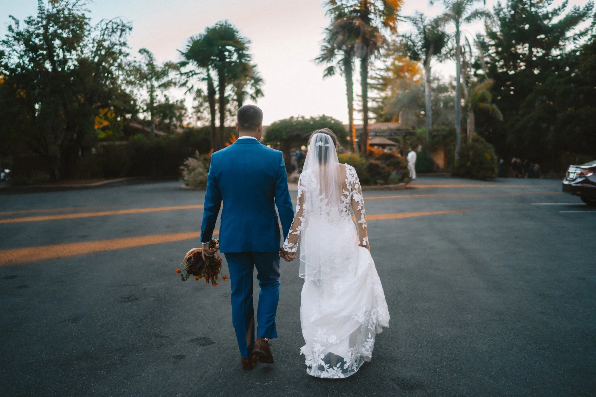 Bride and groom walking hand in hand on a paved road at sunset, surrounded by trees and parked cars.
