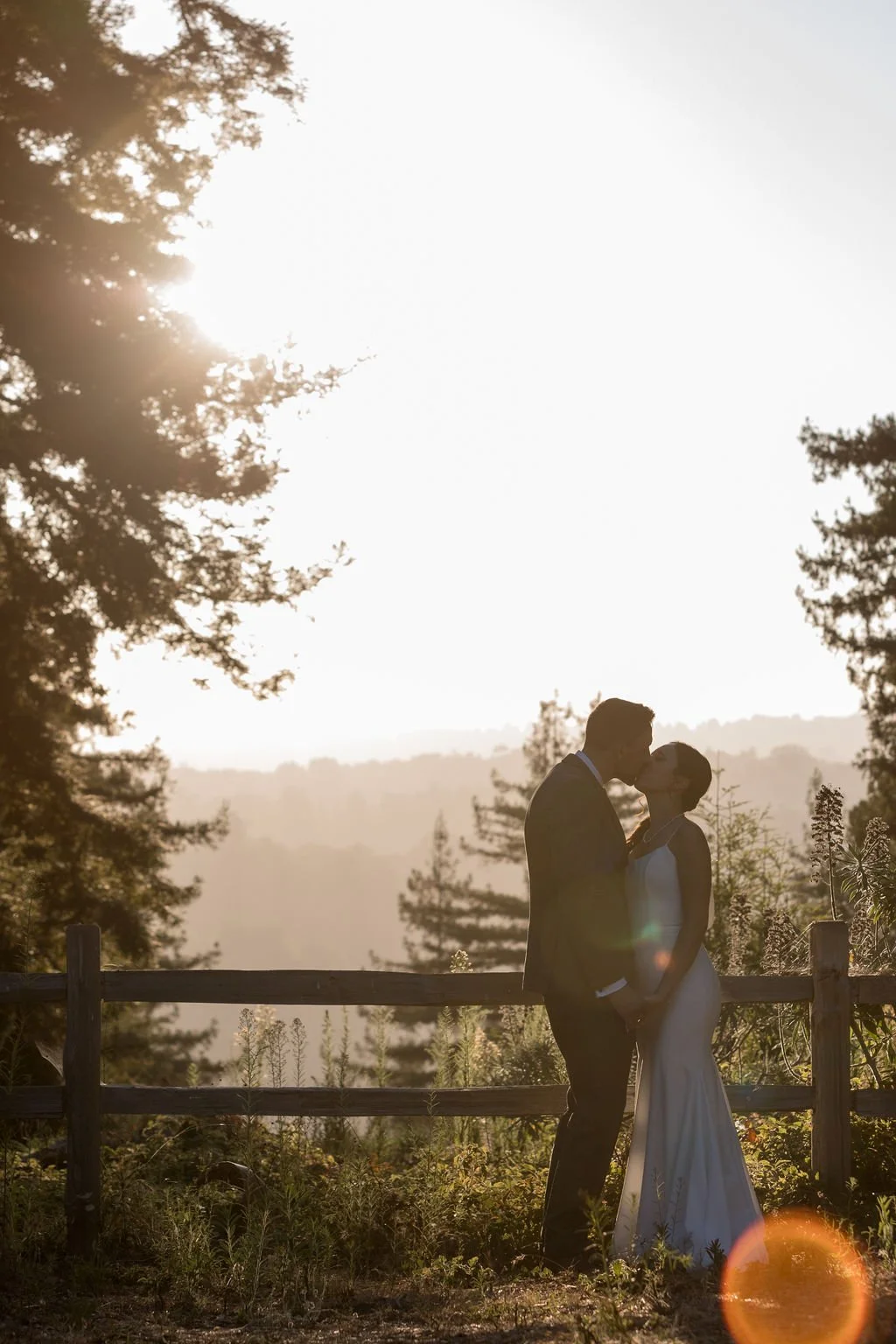 A couple in wedding attire sharing a kiss outdoors at sunset, surrounded by trees and a wooden fence.
