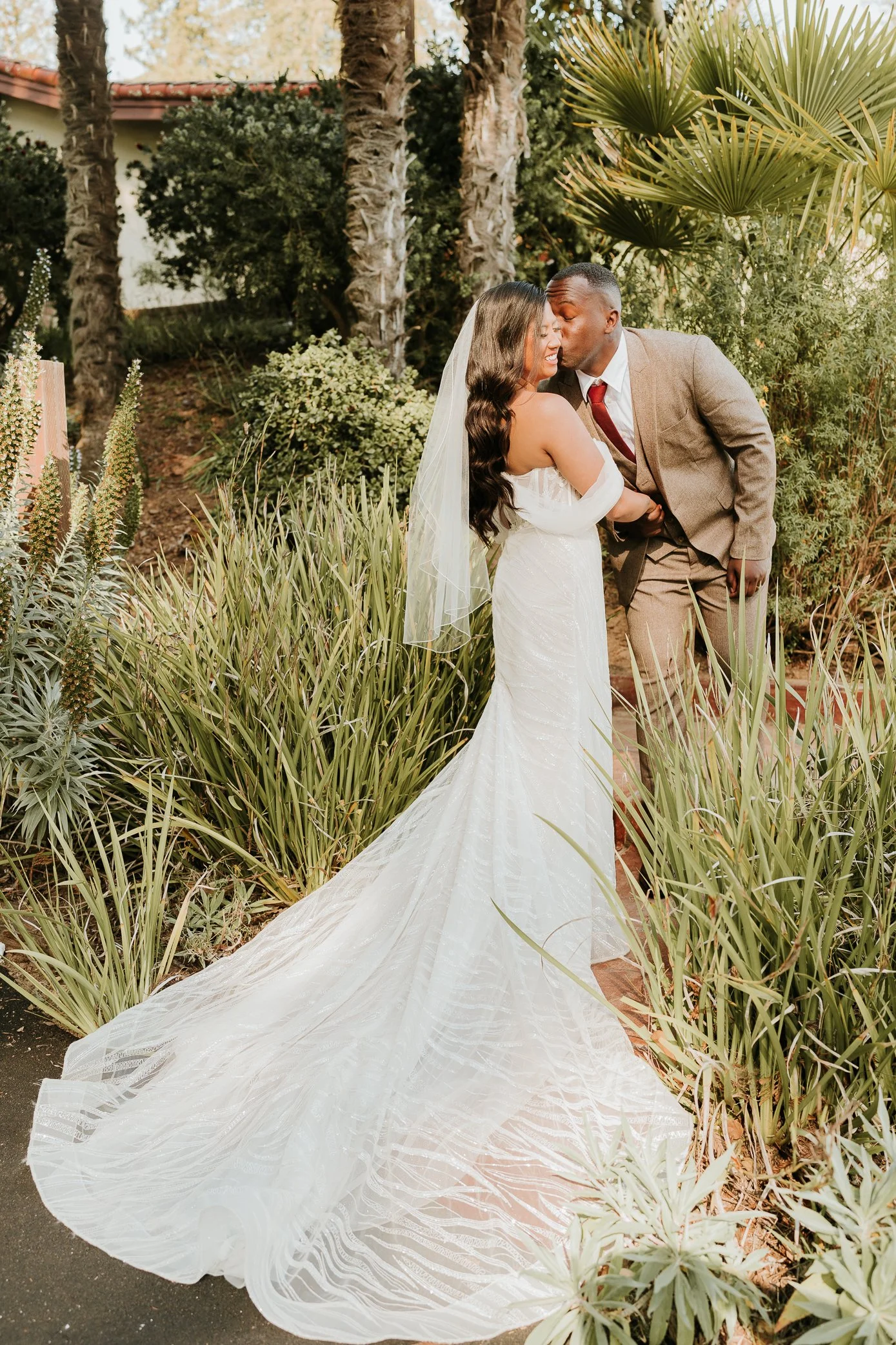 A bride and groom share a tender moment outdoors surrounded by tall plants and palm trees. The bride wears a white wedding gown with a train and veil, and the groom is dressed in a brown suit with a red tie.