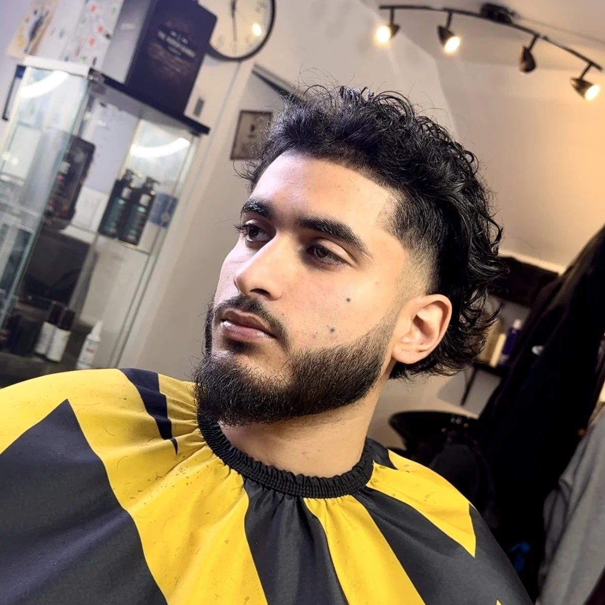 A man with a styled, curly mullet haircut and beard sitting in a barber shop, wearing a black and yellow barber cape, looking to the side.