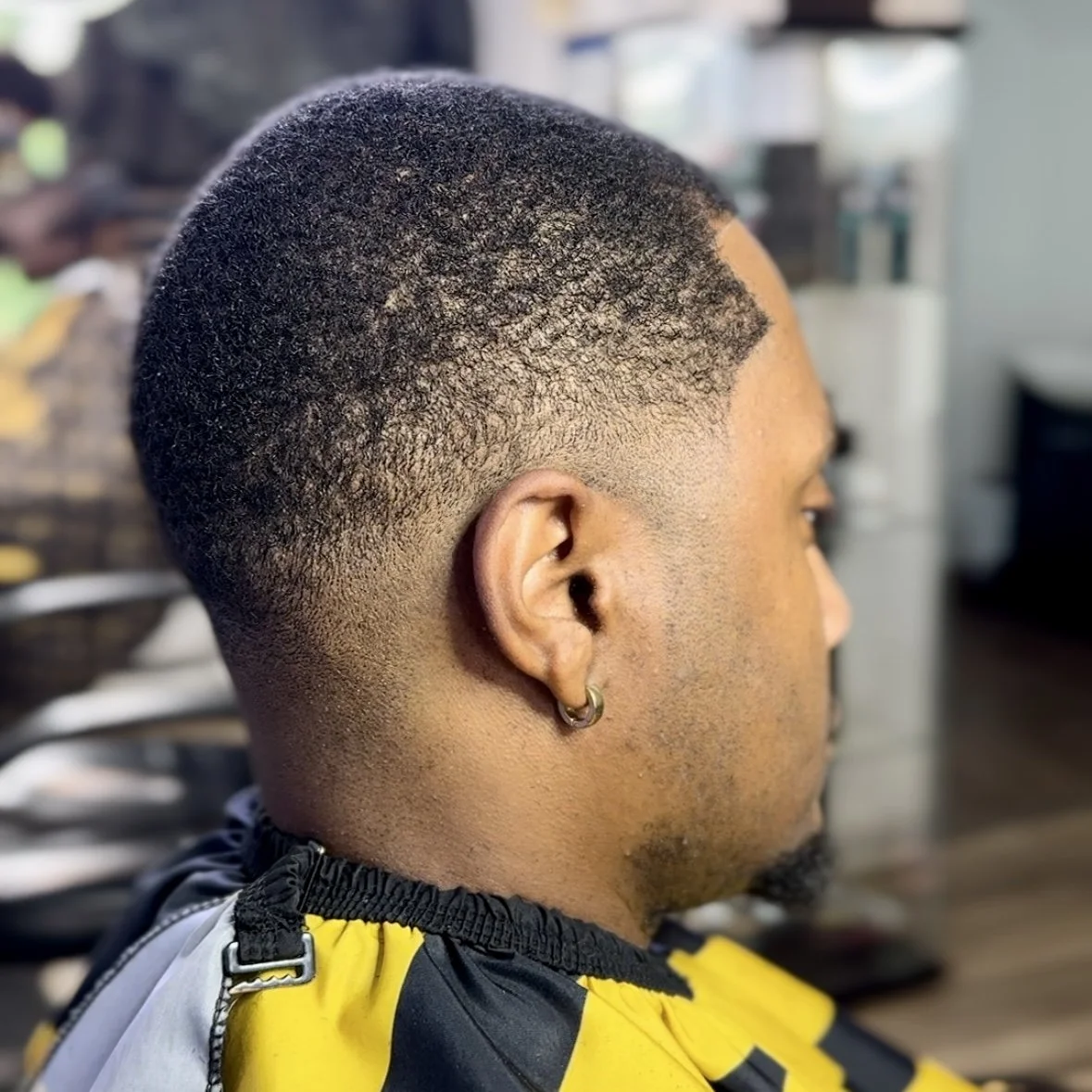Close-up side view of a man with a fresh hairstyle featuring a fade cut, wearing a yellow and black barber cape, with a small hoop earring in his ear, in a barber shop.