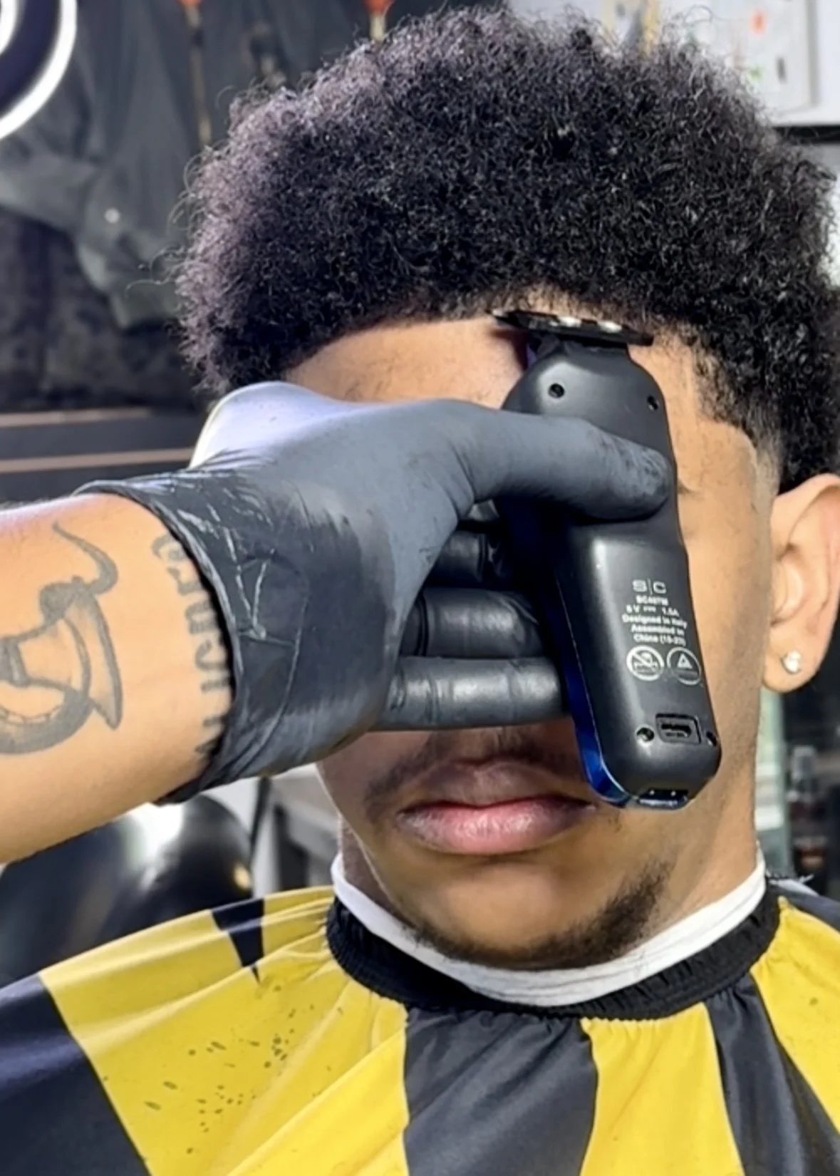 Barber using electric razor to shave customer at barbershop, customer with curly hair wearing a yellow and black barber cape.
