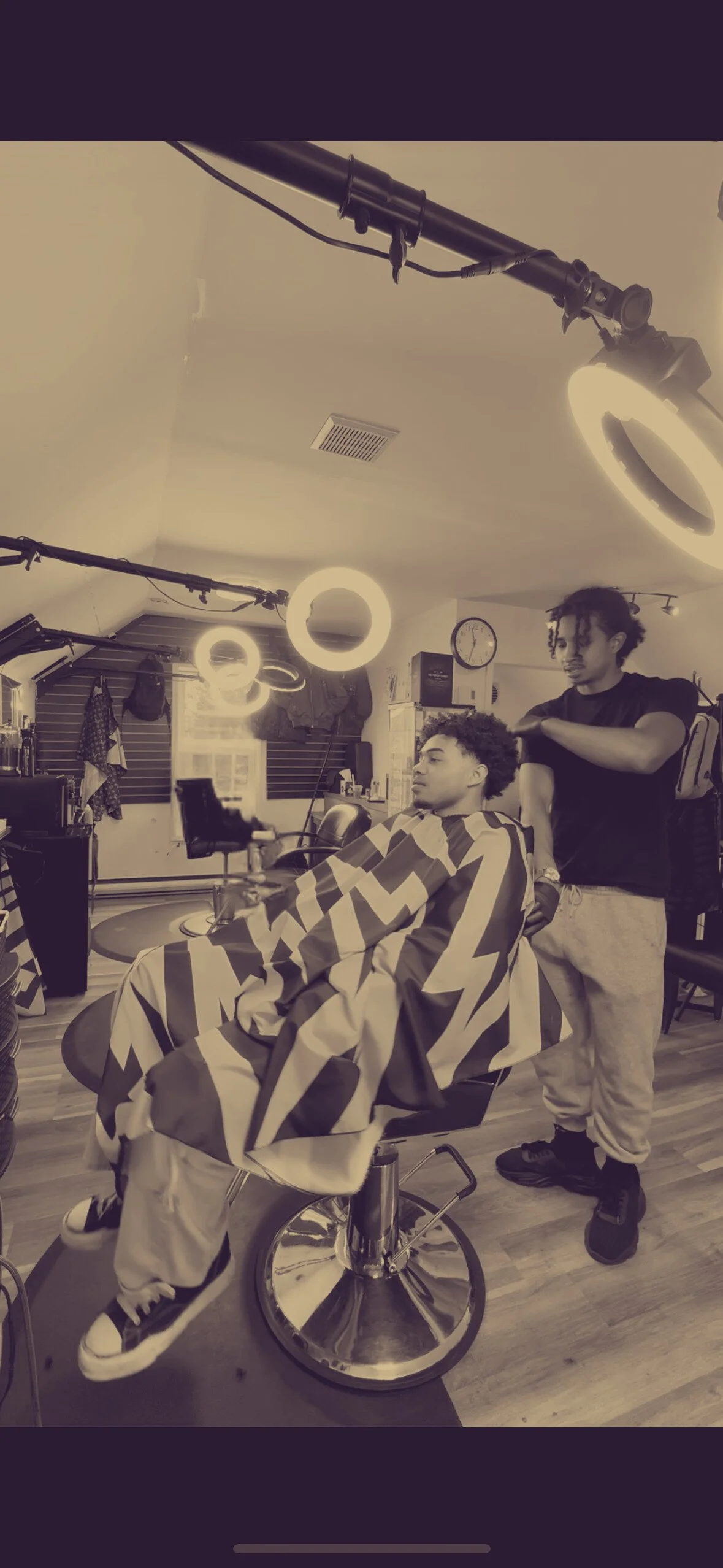 A man getting a haircut at a barbershop, seated in a barber's chair with a geometric-patterned cape, while a barber stands behind him, working on his hair. The shop features ceiling lights, a clock, and various barber tools and supplies.