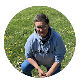 Junge Frau auf grünem Gras mit kleinen gelben Blumen, trägt eine graue Jacke und liest ein Buch.