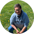 Junge Frau auf grünem Gras mit kleinen gelben Blumen, trägt eine graue Jacke und liest ein Buch.