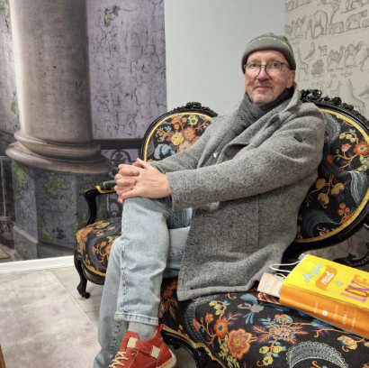 Ein Mann sitzt auf einem gepflanzten, schwarzen Sofa mit buntem Blumenmuster, trägt eine graue Jacke, Mütze und Brille, neben einem Buch und einem Notizbuch.