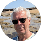 Ein älterer Mann mit weißen Haaren und Sonnenbrille steht an einem Strand oder mineralhaltigen See, im Hintergrund Sand und Wasser.