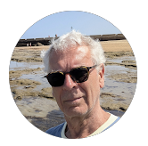 Ein älterer Mann mit weißen Haaren und Sonnenbrille steht an einem Strand oder mineralhaltigen See, im Hintergrund Sand und Wasser.