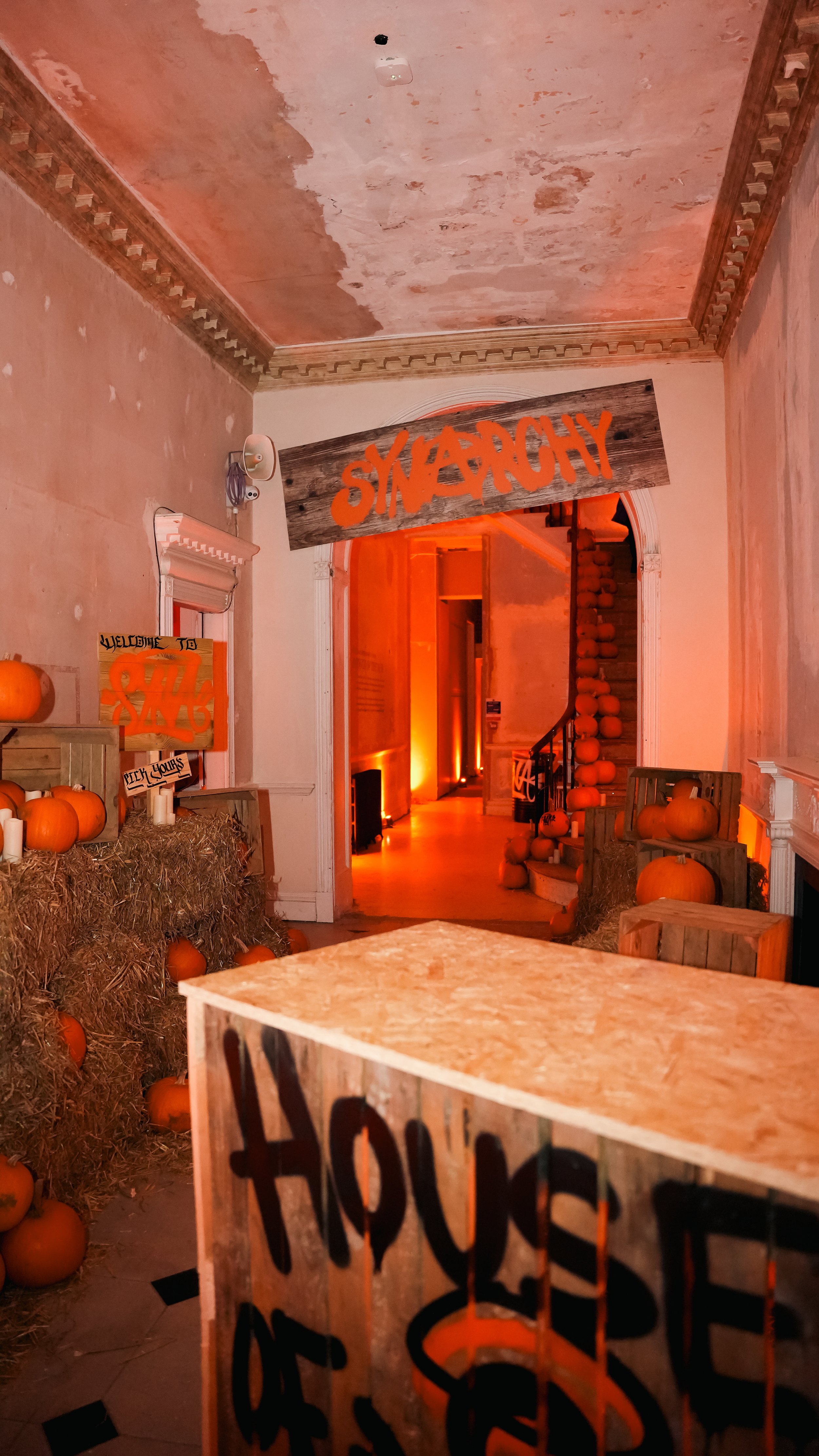 Interior hallway decorated for Halloween with pumpkins, hay bales, and graffiti-style signs, featuring an orange-lit corridor and "SYNARCHY" written above an archway.