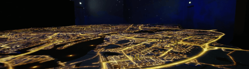 A model of a city at night with illuminated streets and a river running through it, viewed from above.