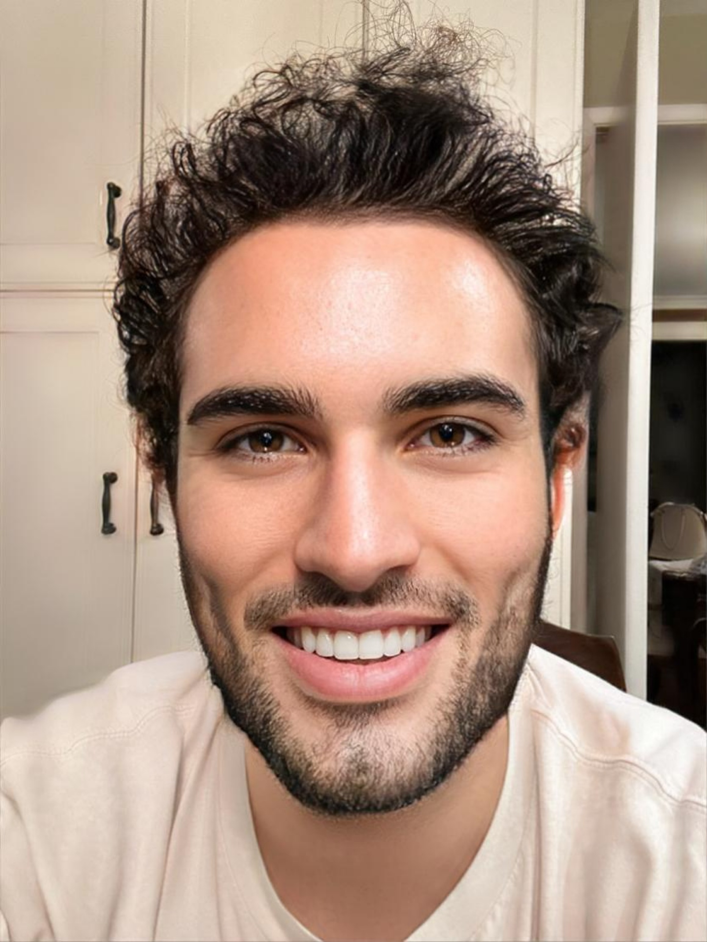 Close-up of a smiling young man with dark curly hair and a beard, indoors with beige cabinets and shelves in the background.