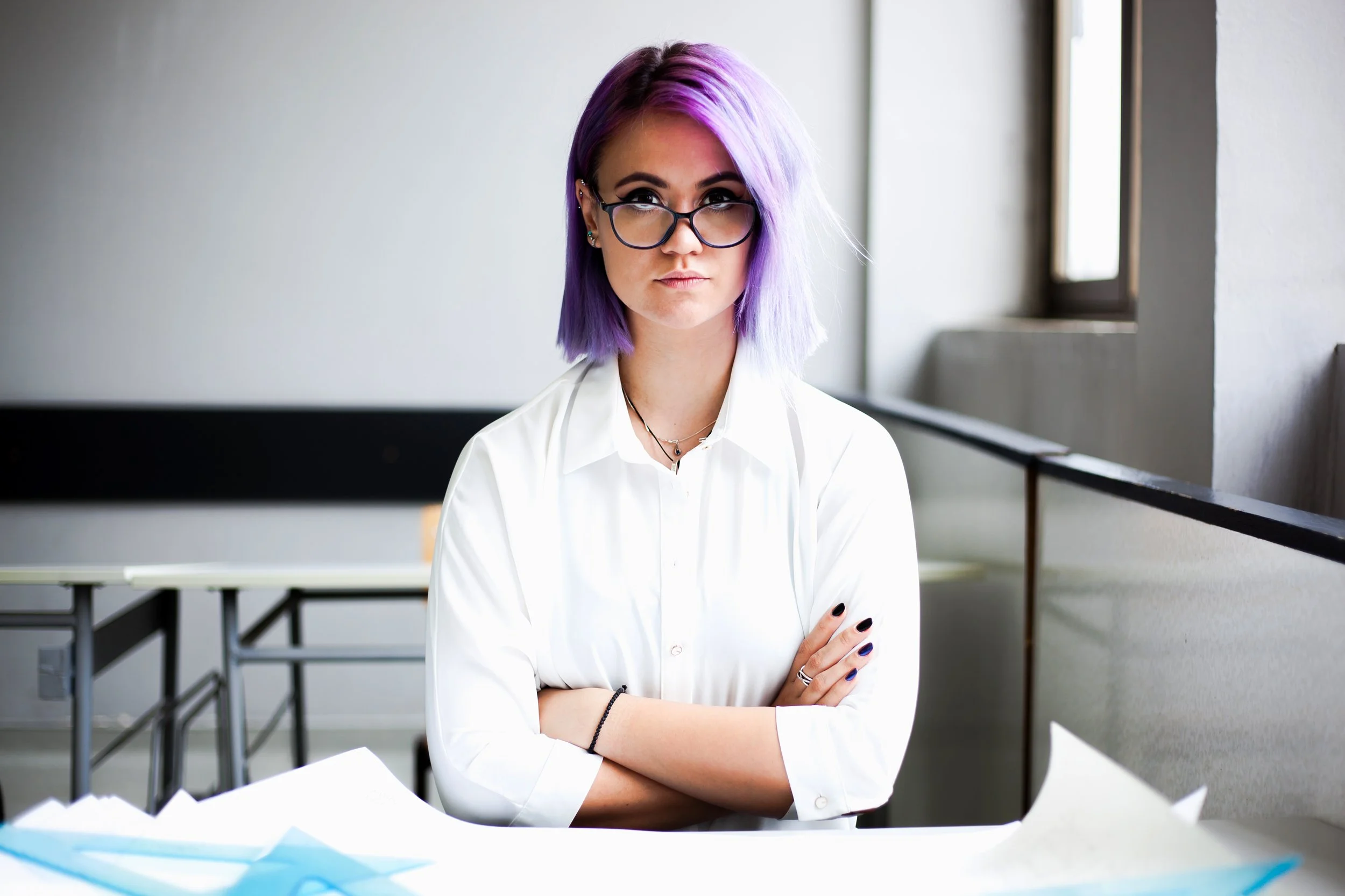 A young woman with purple hair, black glasses, and multiple earrings, sitting with crossed arms in a brightly lit room with windows and a table.