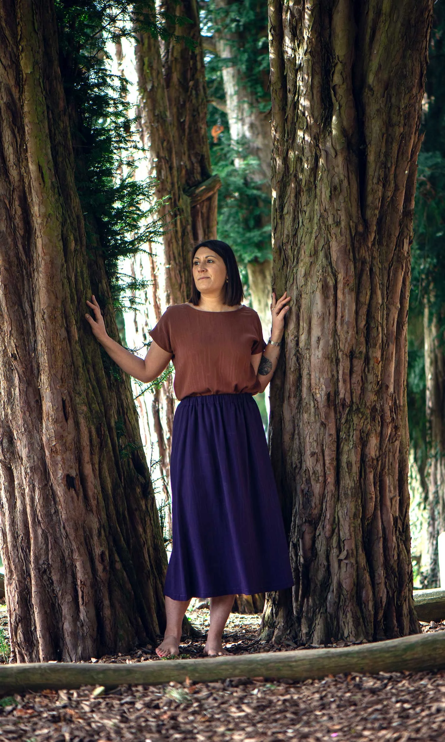 Femme portant une chemise marron et une longue jupe violette, debout entre de grands arbres dans une forêt, touchant deux troncs d'arbre.