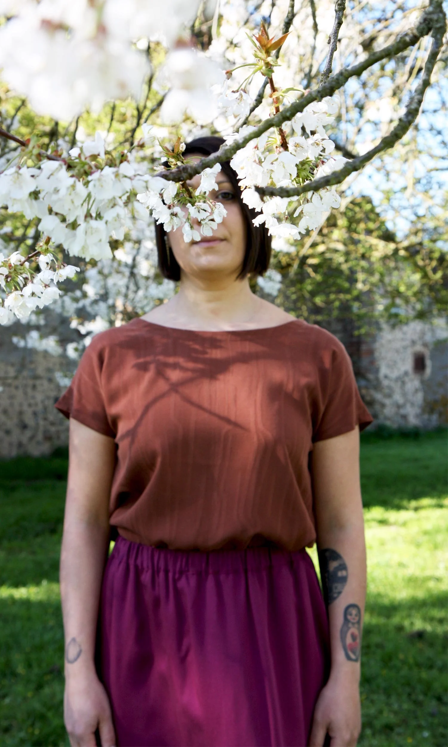 Une femme debout dans un jardin, partiellement cachée derrière une branche de fleurs blanches en fleurs, portant une robe marron avec une jupe pourpre, avec des tatouages visibles sur ses bras, en arrière-plan un arbre et un mur.