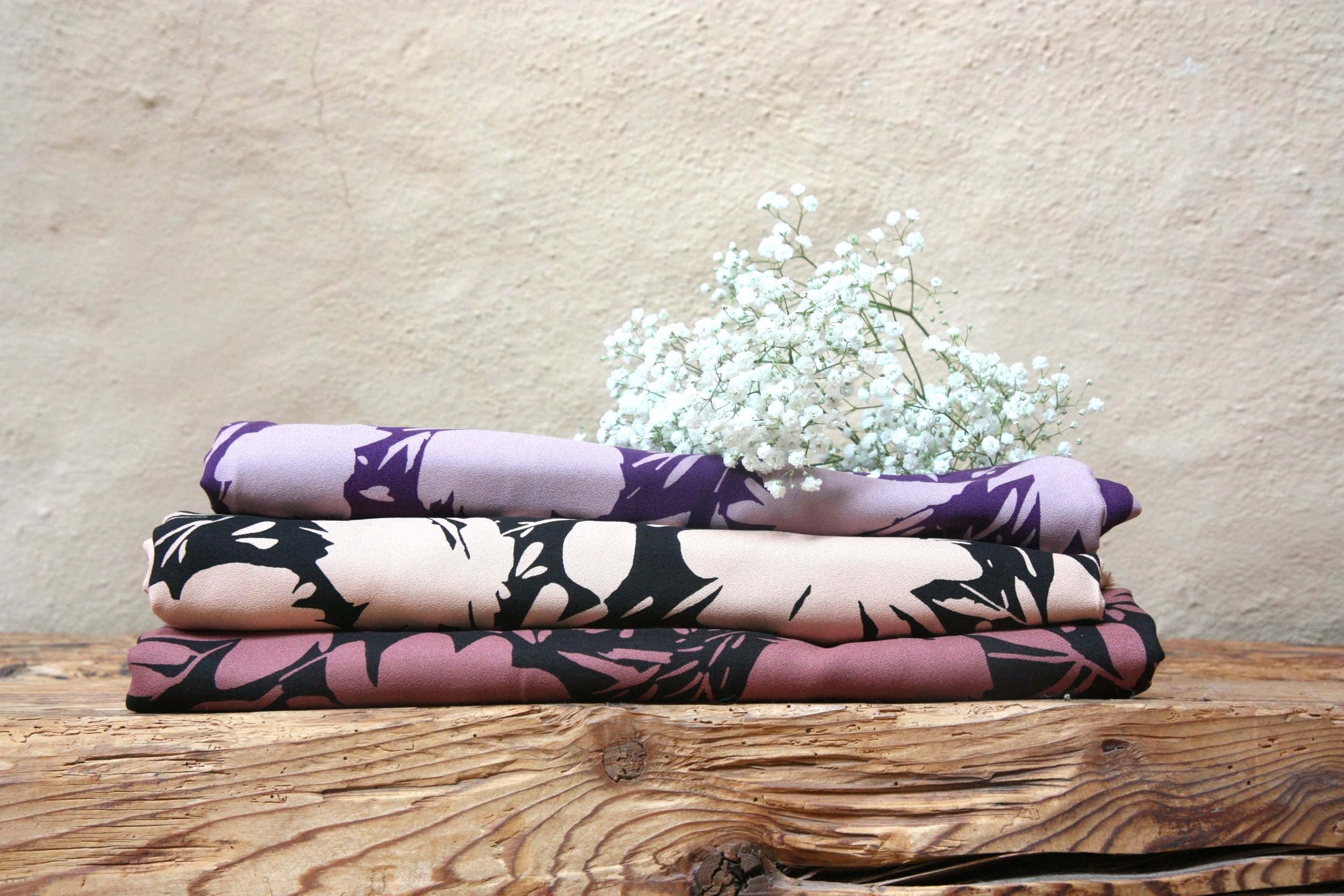 Trois tissus colorés avec des motifs de feuilles, posés en pile sur une table en bois, avec des fleurs blanches en arrière-plan.