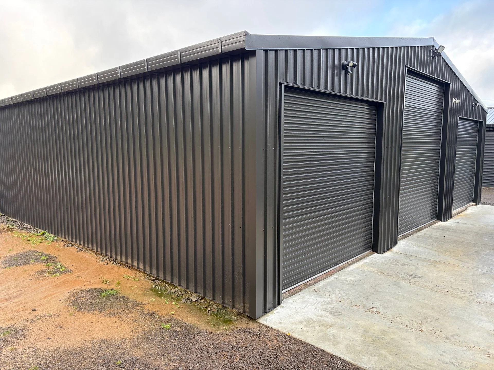 Black metal storage shed featuring durable metal wall cladding, rolling doors and exterior security cameras, set on a concrete pad beside a grass and dirt area.