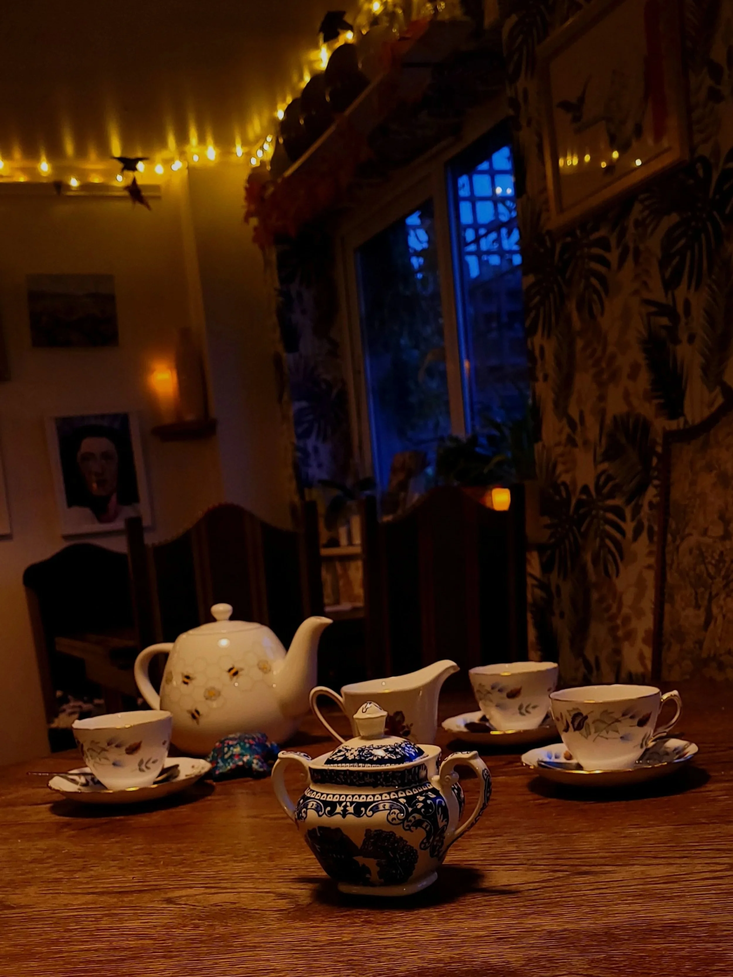 Vintage tea set with a teapot, a sugar bowl, and three matching cups and saucers on a wooden table in a cozy room with fairy lights. The leftovers from afternoon tea in Walthamstow's secret café.