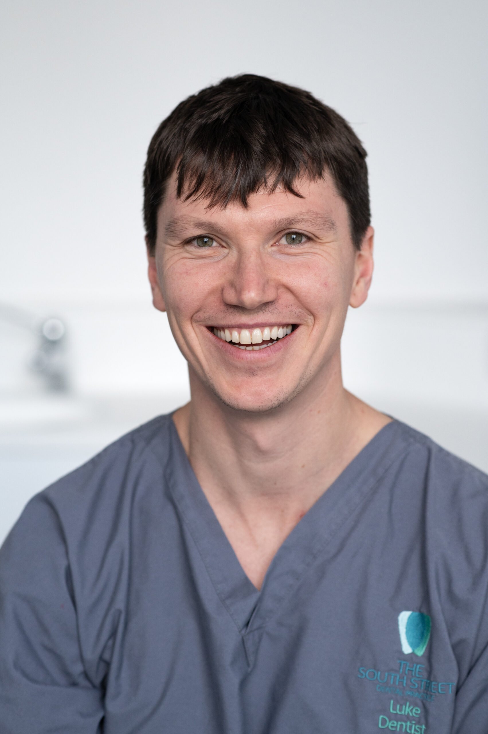 A smiling man with short dark hair wearing gray scrubs with the logo of The South Street Clinic and the name Luke Dentist embroidered on it. The background is plain and blurred.