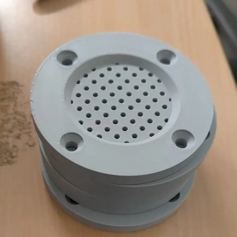 Een ronde, grijs kunststof object wat dient als zwembad filter met tientallen kleine gaatjes in het midden, vier gaten aan de rand, en een zwenkarm rondom. Het ligt op een houten oppervlak.