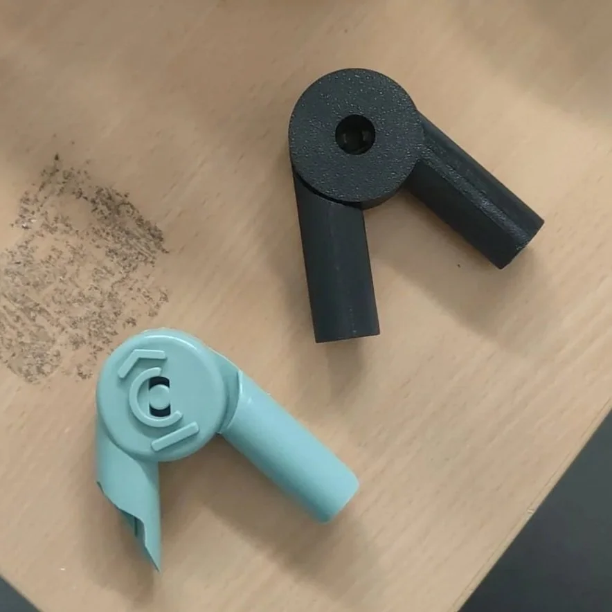 Twee kunststof scharnieren, een zwarte en een lichtblauwe, liggen op een houten tafel. De zwarte is 3D geprint omdat de originele is gebroken.