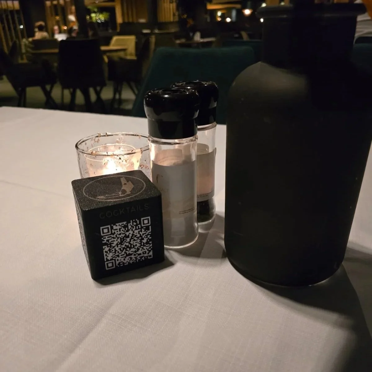Een gedekte tafel in een restaurant 's avonds met een QR-code blok van Van der Valk, zout- en peperstellen, een kaars en een grote donker gekleurde fles op een witte tafel. In de achtergrond zijn andere tafels en stoelen te zien.
