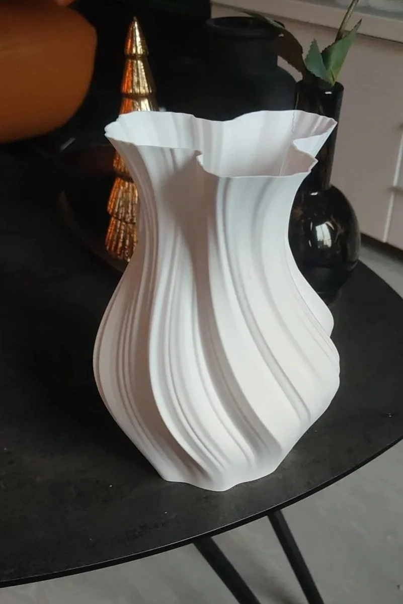 Een modern witte kunststof vaas met een golvend patroon op een zwarte tafel, met decoratieve objecten op de achtergrond.