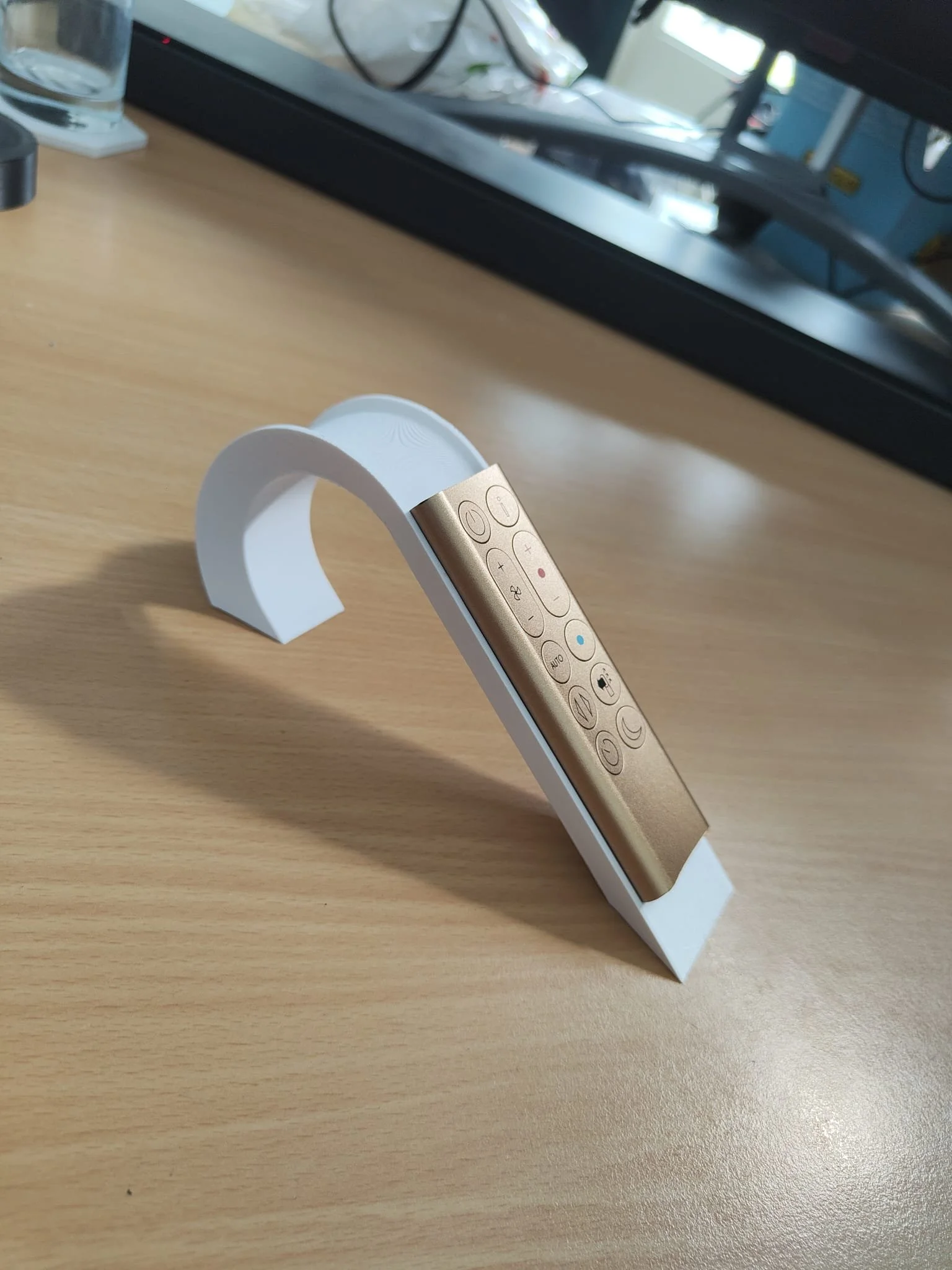 Een afstandsbediening met een witte houder op een houten bureau gemaakt voor Dyson airpurifiers, met een computermonitor en andere kantoorbenodigdheden op de achtergrond.