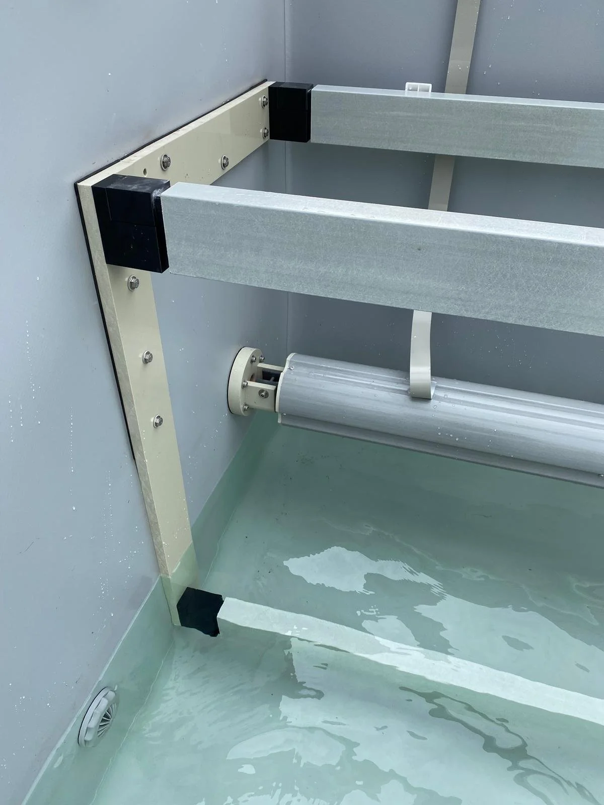 Onderdelen van een mechanisch systeem met metalen en kunststof onderdelen en een cilinder, bevestigd aan een muur van een zwembad met water op de vloer.