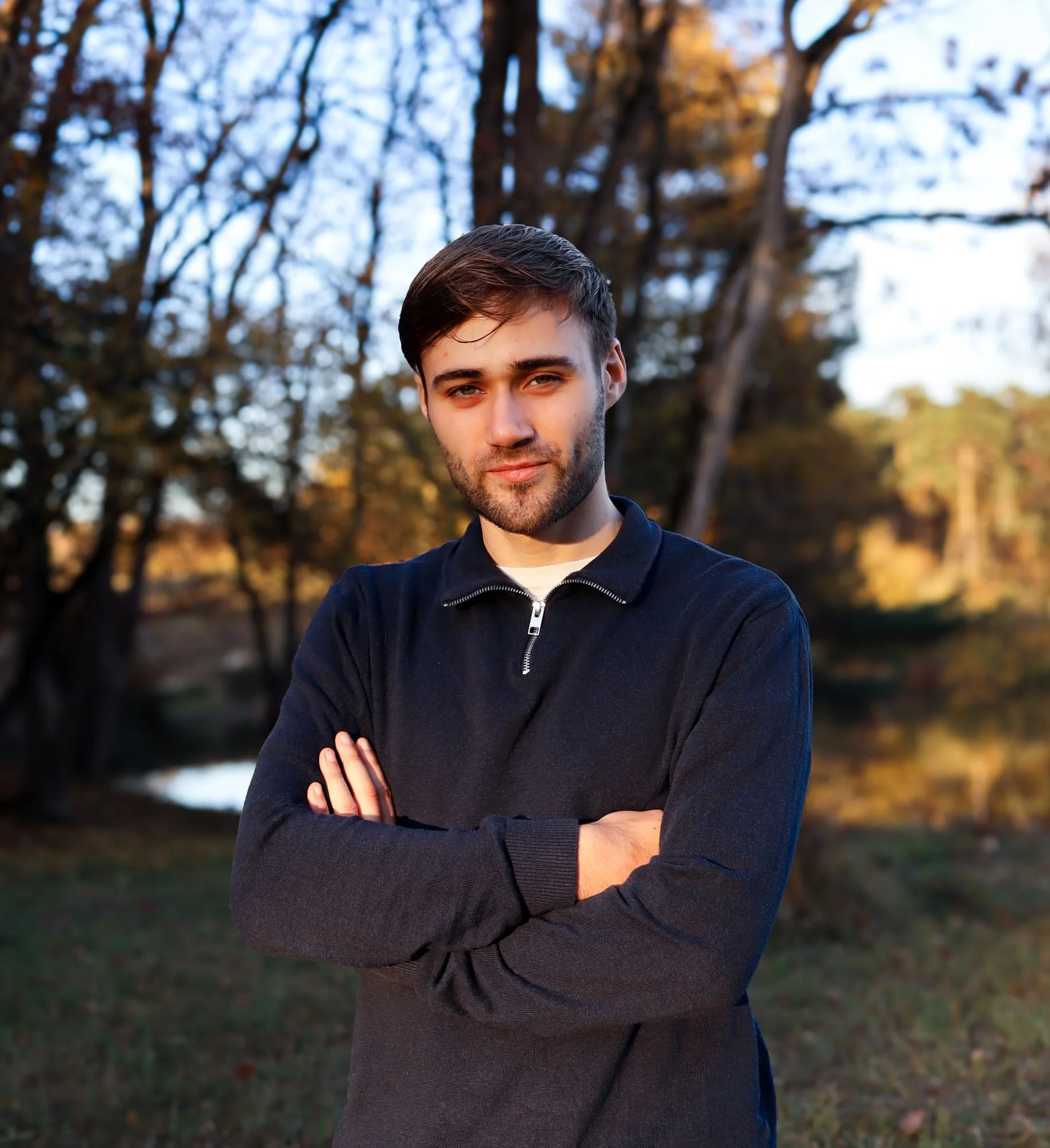 een jonge man genaamd Luuk Wirken met een donkere trui en een lichte shirt eronder, met gevouwen armen, staat buiten tijdens de schemering met bomen op de achtergrond.