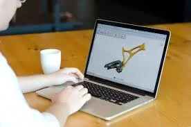 Persoon die op een laptop werkt met een 3D-model op het scherm, op een houten tafel met een witte beker.