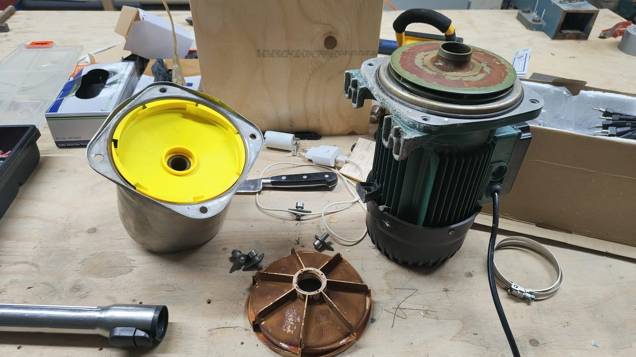 Een werkbank met daarop een waterpomp, een roestige ventilatorkap en verschillende gereedschappen en onderdelen. Het gele onderdeel is een 3D geprinte vervanging van de originele centrifuge kap. 